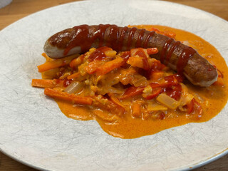 eine wurst auf rotem curry auf einem weissen teller. auf der wurst ist noch etwas siracha zu sehen