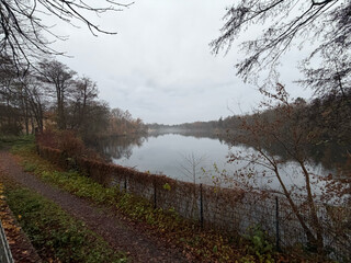 weitwinkelfoto vom pl&ouml;tzensee, aufgenommen am s&uuml;dufer. es ist diesig und grau, der see ganz ruhig, im hintergrund sieht man wasserv&ouml;gel.