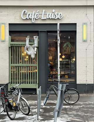 foto vom eingang des caf&eacute; louise. die bank vor dem eingang ist eine skulptur, eine realistische, weiss angemaclte schaumstofffigur sitzt kopf&uuml;ber auf der bank in 3 meter h&ouml;he