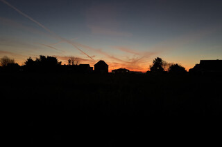 sonnenaufgang in karken mit kahlen b&auml;umen und haus-silhouetten am horizont. der himmel leuchtet in warmen orange- und gelbt&ouml;nen.