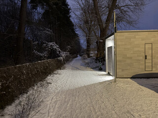 foto einer &ouml;ffentlichen toilette im fr&uuml;hen morgengrauen. das licht aus den beiden offenen pissoir-kabinen scheint einladend auf einen schneebedeckten hohlweg